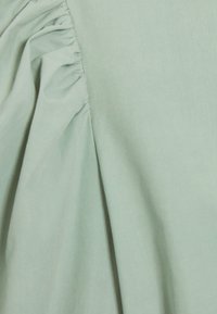 Světlý zelený materiál s nařasenými záhyby, hladkou texturou a jemným leskem. Design obsahuje měkký efekt splývání na jedné straně.