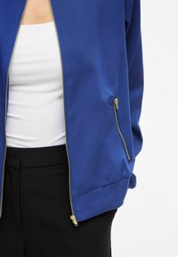 Une personne portant une veste bleue zippée par-dessus un haut blanc et un pantalon noir, avec la veste partiellement ouverte et les mains dans les poches.