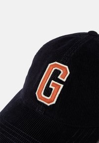 Cappello in corduroy blu navy caratterizzato da una "G" ricamata in arancione e bianco, con evidenti striature di trama nel tessuto.