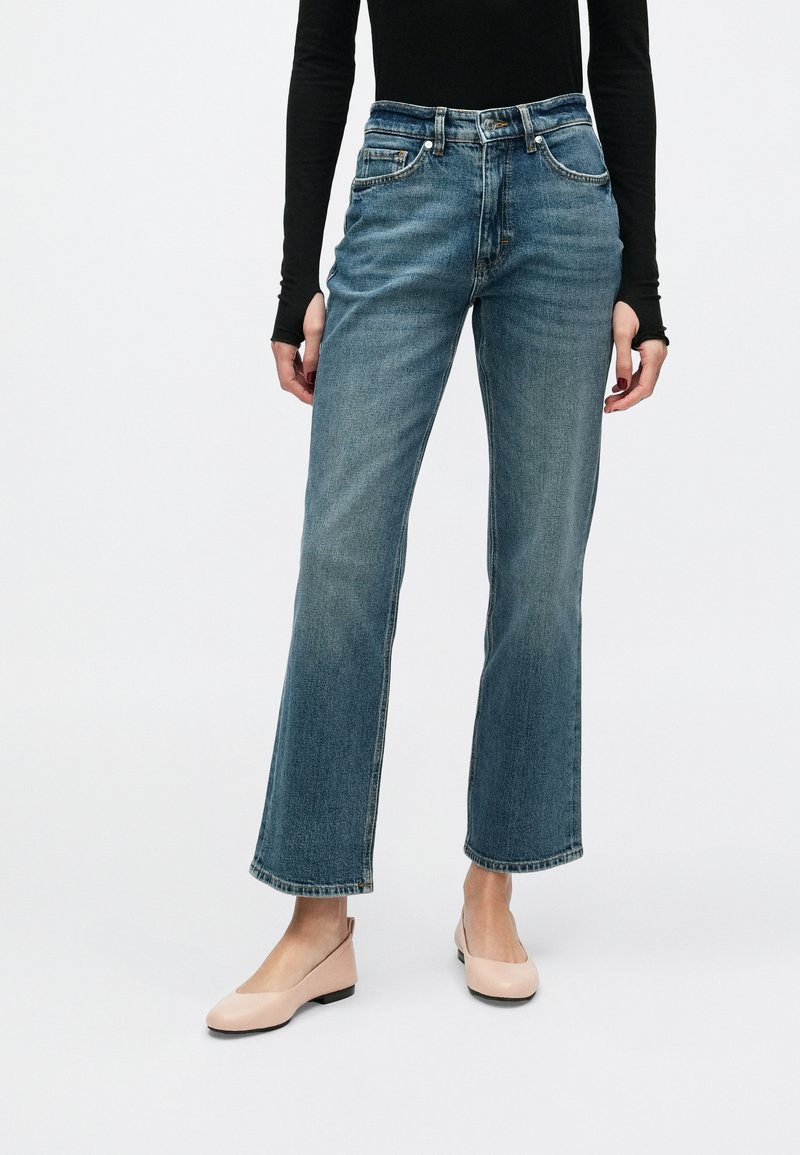 Kék farmernadrág, magas derékkal, egyenes szárú kivitelben, kopott hatású részletekkel és ötpoharas kialakítással, világos rózsaszín lapos cipőkkel párosítva.