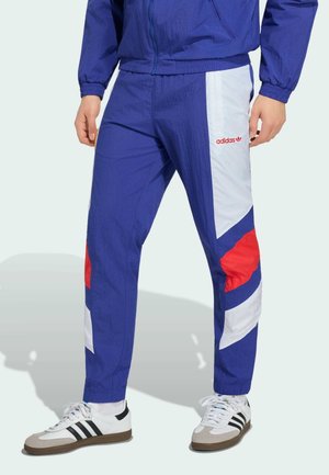 Mężczyzna w niebieskich, białych i czerwonych spodniach dresowych Adidas oraz białych butach Adidas z czarnymi paskami, stoi z rozluźnionymi rękami.