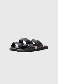 Sandales bicolores avec un design en cuir noir et marron, à bout ouvert, deux brides avec des boucles métalliques, et une semelle plate en caoutchouc.