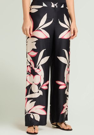 Kobieta w czarnych szerokich spodniach z dużym beżowo-różowym kwiatowym wzorem oraz czarnymi sandałami ze złotymi akcentami.