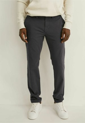 Mężczyzna ubrany w ciemnoszare spodnie o prostych nogawkach, białe trampki i kremową bluzę z długim rękawem, stoi na jasnym tle.