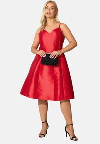 Vestido rojo de satén en corte A con escote en forma de corazón, tirantes y falda acampanada. Los accesorios incluyen un bolso de mano negro con flecos y tacones negros.