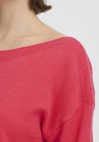 Pull en tricot rose avec un large col et des bords côtelés, présentant une texture douce et une coupe décontractée. Vue rapprochée de la zone de l'épaule.