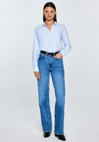 Sieviete stāv, valkājot gaismas zilu pogājamu kreklu, zilas augstas jostasvietas džinsas ar melnu jostu un melnas potīšu zābaciņus, rokas atslābinātas vai kabatās.