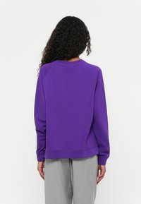 Felpa viola con scollo rotondo e maniche raglan, realizzata in tessuto morbido. L'orlo si trova all'altezza della vita, abbinata a pantaloni a righe grigio chiaro.