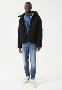 Chaqueta acolchada negra con capucha, cierre de cremallera y bolsillos laterales, combinada con una camisa de mezclilla clara y jeans azules ligeramente desgastados.