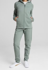 Lichtgroene hoodie met rits en zakken, gecombineerd met bijpassende broek, gemaakt van zacht materiaal. Gecombineerd met witte sneakers met een gouden accent.