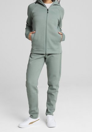 Sudadera con capucha verde claro con cremallera y bolsillos, junto con pantalones a juego, hechos de un material suave. Combinado con zapatillas blancas con un detalle dorado.