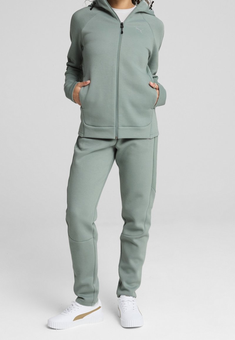 Lichtgroene hoodie met rits en zakken, gecombineerd met bijpassende broek, gemaakt van zacht materiaal. Gecombineerd met witte sneakers met een gouden accent.