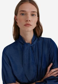 Donkerblauwe blouse van gladde stof, met een vastgebonden kraag en lange mouwen. Het ontwerp heeft een relaxte pasvorm en een minimalistische textuur.