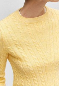 Keltainen palmikkoneulepaita, jossa on pyöreä pääntie ja teksturoitu kuvio. Kangas näyttää pehmeältä, kevyeltä ja hieman istuvalta.