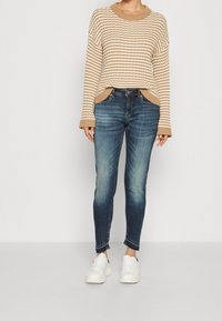 Suéter a rayas en tonos beige y crema con mangas anchas, combinado con jeans oscuros de corte slim y zapatillas blancas. Los jeans tienen los bajos deshilachados.