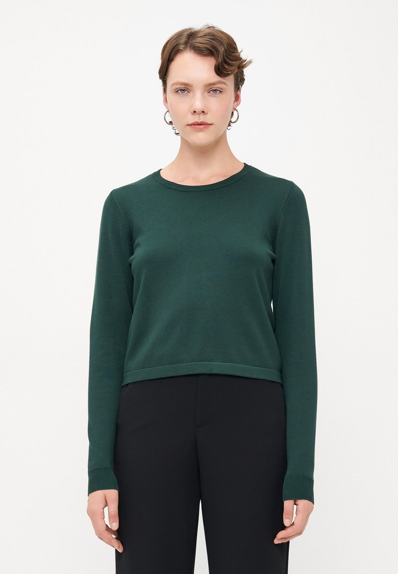 Pull en maille vert avec des manches longues, un col rond et un bord-côte aux poignets et à l'ourlet. Porté sur un pantalon noir. Texture lisse.
