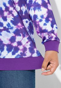 Felpa a tinta unita viola e blu con maniche lunghe e polsini a costine. L'orlo è anch'esso a costine, realizzato in un tessuto liscio e dalla texture. 
