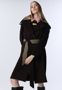 Jonge vrouw in een donkerbruine gewatteerde jas met een riem en geruite details, met een roze overhemd met kraag, poseert tegen een lichtgrijze achtergrond.