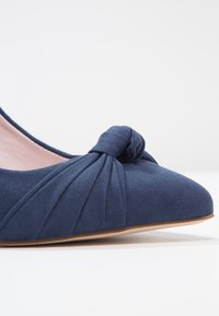 Chaussure plate bleu marine avec un détail de nœud noué sur le bout, présentant une texture douce en suédine et un design de bout pointu et arrondi.