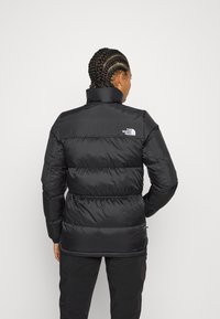 Black puffer jacket with a high collar, quilted design, and elastic cuffs; features a small logo on the upper back. Smooth texture.