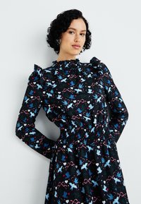 Vestido preto com mangas longas, detalhes em babados, cintura ajustada com pregas, e padrões coloridos de pássaros e corações por toda a peça.