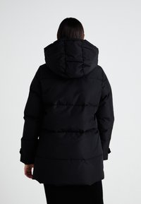 Veste doudoune noire avec capuche, design matelassé et manches longues. Le tissu semble lisse et est conçu pour la chaleur.