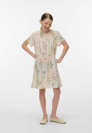 Ragazza in piedi con le mani sui fianchi, indossa un vestito floreale color crema e scarpe piatte beige intrecciate, sorride con gli occhi chiusi su sfondo bianco.