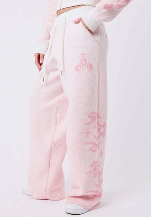 Femme portant un pantalon de jogging ample rose clair avec cordons et motifs roses plus foncés subtils, debout avec une main dans la poche et des baskets blanches.