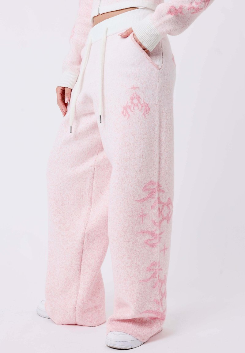 Woman wearing light pink wide-leg sweatpants with drawstrings and subtle darker pink patterns, standing with one hand in pocket and white sneakers.