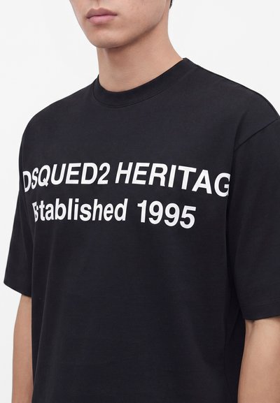 Νεαρό άτομο που φοράει μαύρο T-shirt με λευκό κείμενο "DSQUED2 HERITAG Established 1995" στο μπροστινό μέρος.