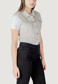Gilet di jeans grigio chiaro tagliato con chiusura a bottoni, colletto a punta e tasche sul petto, indossato sopra una maglietta bianca e abbinato a jeans neri.