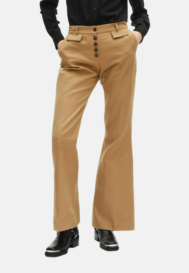 Tan wijde pijpen broek van een gladde stof, met een hoge taille, zijzakken en zes donkere knopen aan de voorkant. Gemaakt in combinatie met zwarte laarzen.