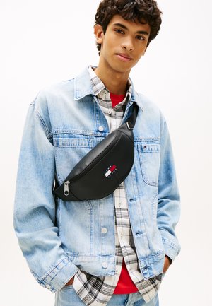 Sort læder bæltetaske med lynlås foran, med en logopatch, båret over en denimjakke lagdelt med en ternet skjorte og en rød skjorte.