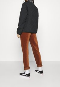 Sort quiltet jakke med et logo på kraven, brune corduroy bukser og sorte Vans sneakers med hvide såler. Hvide sokker synlige.