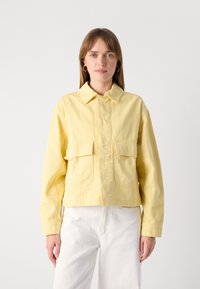 Gul denimjakke med krage, to lommer foran og cropped design. Glatt tekstur og synlige sømdetaljer.