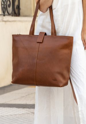 Donna che tiene una grande borsa a tracolla in pelle marrone con manici, indossa un vestito bianco con spacco laterale, su un sentiero esterno.