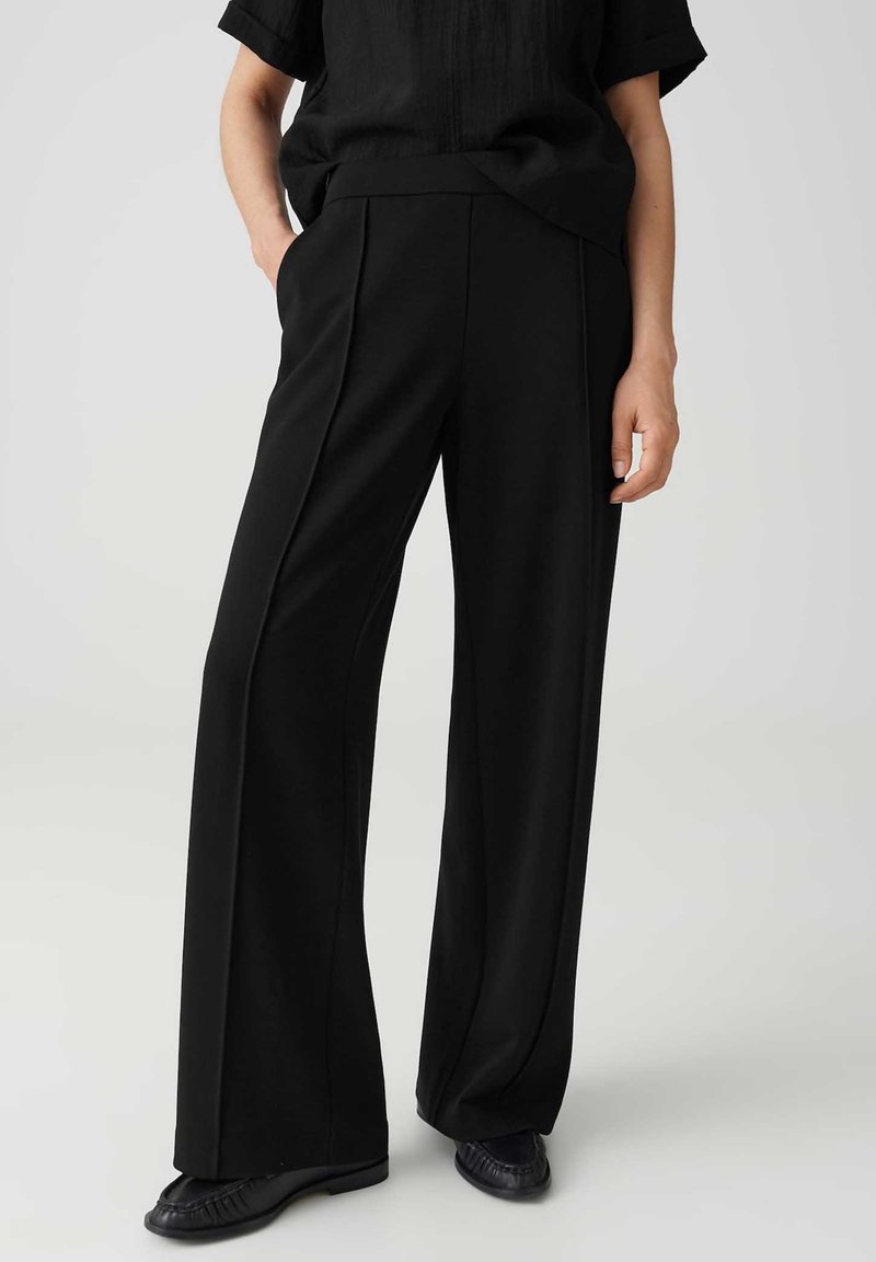 Person wearing black wide-leg trousers and black shoes, standing with one hand in pocket against a plain background.