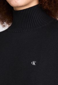 Melnas augstās apkakles džemperis ar ribota raksta virsmu, kuram uz krūtīm ir mazs balts "CK" logotips. Tuvplāns izceļ adīšanas detaļas.