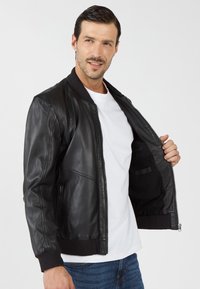Zwarte leren bomberjack met ribgebreide cuffs en kraag, voorzien van een ritssluiting aan de voorkant en binnenzakken, gedeeltelijk open weergegeven.