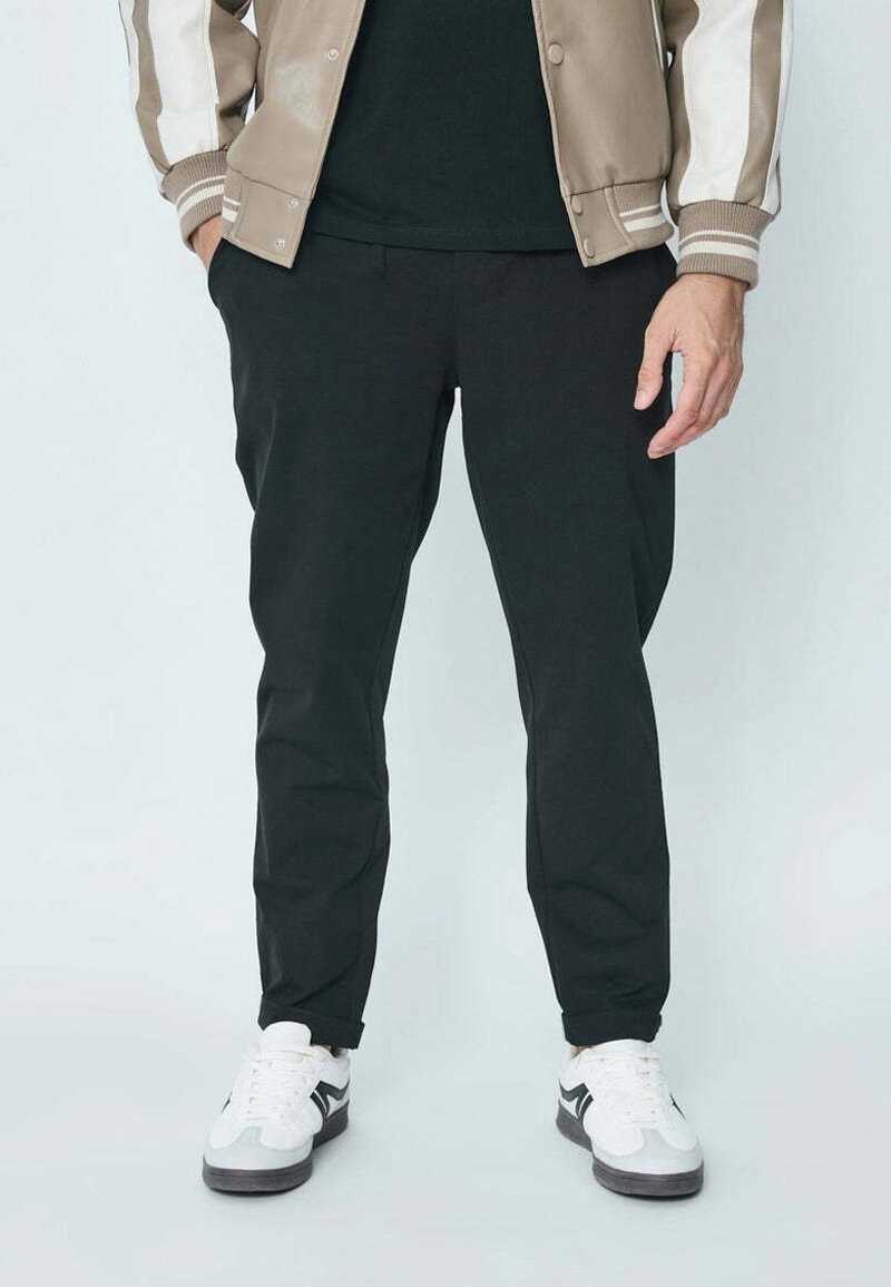 Pantalons de jogging noirs fuselés en tissu doux, associés à des chaussures de sport blanches et grises. La tenue comprend une veste bomber beige avec des rayures blanches.