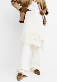 Gonna a portafoglio crema con pizzo, abbinata a pantaloni bianchi a gamba larga e scarpe in suede marrone. Look stratificato con un giubbotto marrone neutro.