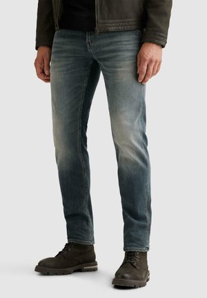 Homme portant un jean bleu délavé, une veste en cuir noire et des bottes marron foncé à lacets, debout devant un fond uni.