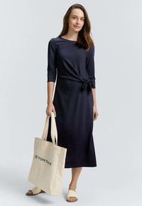 Tumši zila, ceļgalu garuma kleita ar jostu viduklī. Modelis tur bēša krāsas somu ar "TOM TAILOR" logotipu. Vienkāršs dizains un mīksts audums.