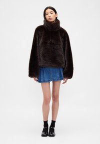 Pullover marrone in pelliccia sintetica con collo alto, maniche larghe e texture liscia, abbinato a una gonna di jeans blu e scarpe nere.