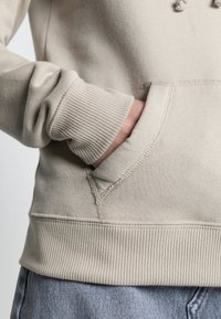 Beige mikina s texturovanou látkou, obsahující klokánek a žebrované manžety a spodní lem. Viditelné švy vylepšují design.