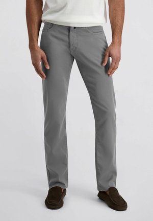 Homme portant un pantalon gris coupe droite, une chemise blanche et des chaussures à enfiler marron foncé, debout devant un fond clair uni.