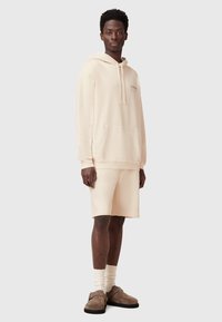 Felpa beige con tasca frontale, pantaloni corti coordinati e calzini leggeri. Tessuto liscio con vestibilità rilassata, caratterizzata da un logo sottile sul petto.