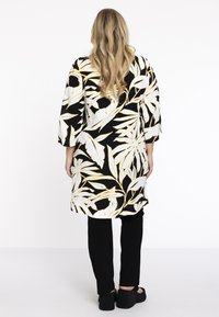 Zwart-witte tuniek met gele bladerenpatronen, driekwart mouwen en een rechte snit, gecombineerd met zwarte broek en zwarte schoenen.