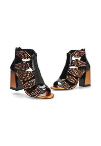 Sandalias de tacón de charol negro con diseño recortado, adornadas con tachuelas moradas y doradas. Tacón ancho, estilo de punta abierta.
