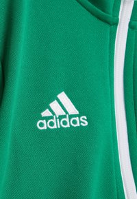 Grøn stofjakke med hvid Adidas-logo broderi og en hvid lynlås, der løber lodret på højre side.